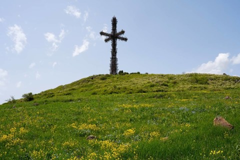 Metal cross on a green hill under a cloudy blue sky, surrounded by grass and yellow flowers,