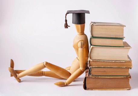 A wooden man in a student cap sits on the floor and leans his back on a stack of textbooks