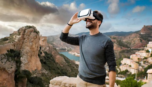 A man wearing a VR headset is standing on a rocky hillside, looking out over a beautiful landscape.