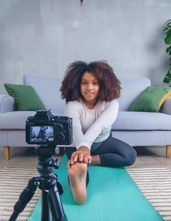 A young woman is doing a yoga pose in front of a camera. The camera is on a tripod and is recording