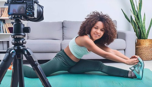 A young woman is stretching on a mat in front of a camera. The camera is on a tripod and is