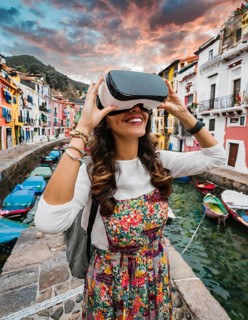 A woman wearing a colorful dress is holding a virtual reality headset. She is smiling and she is