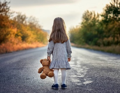 A young child stands alone holding a teddy bear in an urban outdoor setting, symbolizing innocence