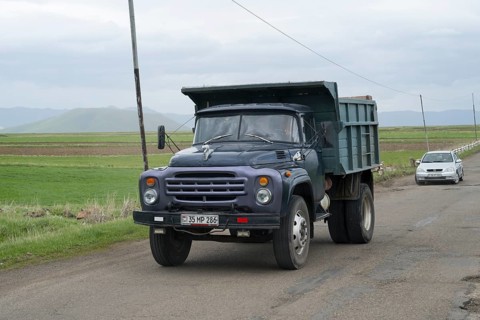 A blue lorry with a trailer drives on a country road surrounded by green fields and mountains,