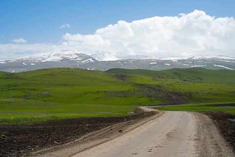 A dirt road winds through green hills in front of a snow-covered mountain range under a cloudy sky,