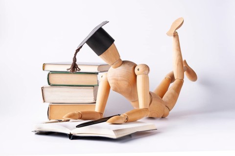 A wooden man in a student hat lies on the floor and reads textbooks