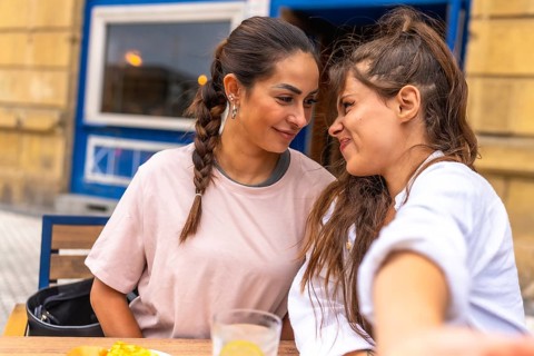 Two women are sitting close together at a table outside a cafe, smiling and enjoying each other's