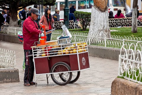 A street vendor operates a fruit stand on a pavement in a busy city, selling fresh juice in the