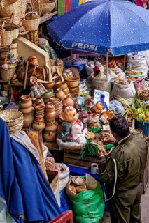 Traders at a lively market with ceramics, dolls and colourful fabrics under a blue umbrella,