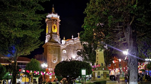 An illuminated church at night decorated with festive activities and flags, surrounded by trees,
