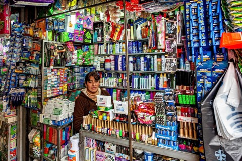 A kiosk with a saleswoman behind the counter, surrounded by a colourful variety of household goods,