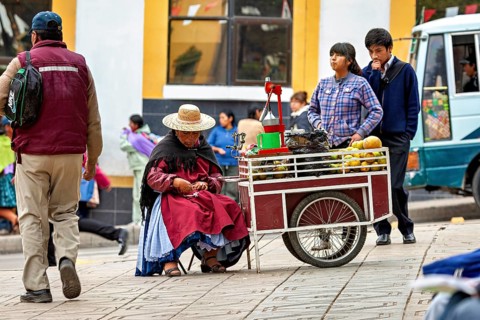 A saleswoman in a hat sits at a fruit stand on a busy pavement, selling fresh juice in the city of