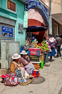 Women in traditional dress sell fruit at a busy market stall in a Bolivian town, selling goods in
