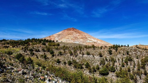 Brown volcanic mountain under a clear blue sky, surrounded by sparse vegetation and trees, the