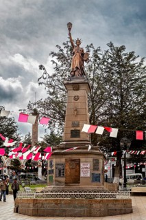 Statue on a stone pedestal in a plaza with red and white flags and trees, under a cloudy sky,