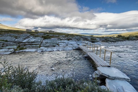 A small wooden bridge crosses a wide river, surrounded by rocks and sparse vegetation in the