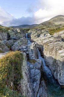 Deep gorge with meandering watercourse, surrounded by jagged rocks and mountains under a cloudy