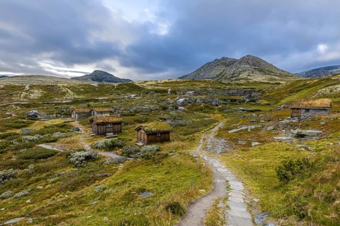 Wooden huts in an open meadow in a remote mountain landscape, with winding paths and clouds in the