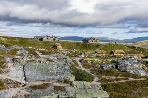 Various wooden cabins on a rocky plain, surrounded by grass and mountains under a cloudy