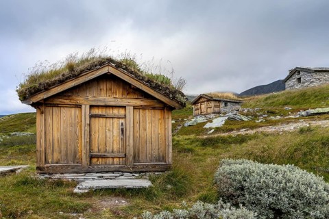 Wooden huts with grass-covered roofs in a hilly, rural landscape under a cloudy sky, Peer Gynt