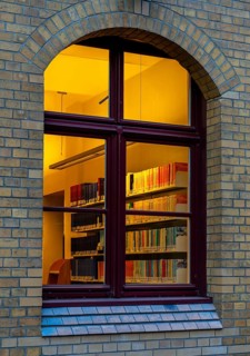 A view through the window of a library, shelves full of books illuminated by a warm, yellow light,