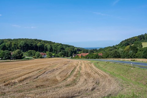 View of a harvested field with neighbouring village and wooded hills under a clear sky, Halle,