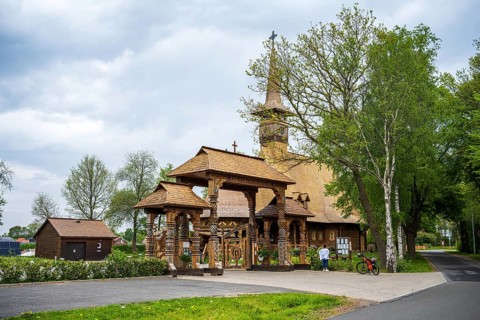 Wooden church with characteristic carvings, along a country road, surrounded by trees and sheds,