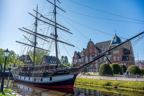 A historic sailing ship on a sunny day with a striking brick building in the background, The