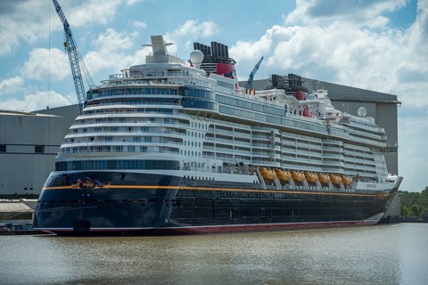 A large cruise ship lies calmly in the water next to a large shipyard on a partly cloudy day,