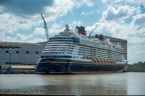 A cruise ship lies in the water in front of a shipyard on a partly cloudy day with a crane in the