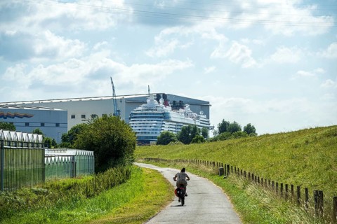 A cyclist on a country road next to a shipyard with a cruise ship in the background, cruise ship at