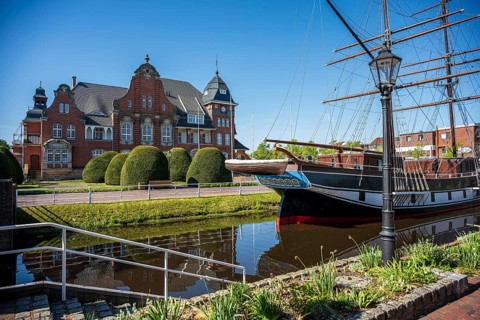 An old ship in front of an impressive brick building, surrounded by manicured nature and water, The