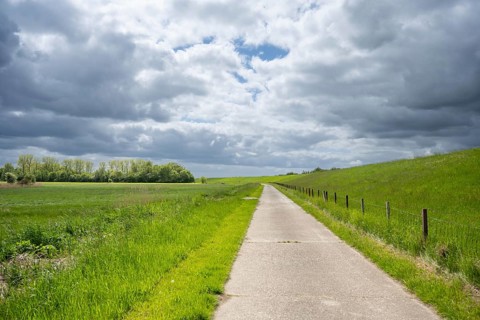 A path leads through a green landscape under a cloudy sky, farm track behind a dyke near
