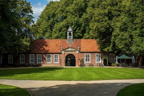 A brick building with a bell tower in front of a grassy area and surrounded by tall trees in the