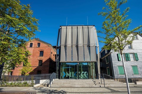 Modern glass building between old houses, surrounded by trees, bright blue sky, Museum of Early