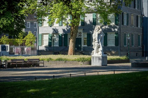 Small square with stone statue in front of an old house with green shutters, illuminated by