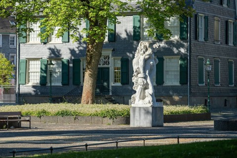 Stone figure in front of a historic building with green shutters, surrounded by blossoming trees,