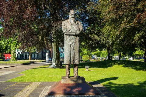 Stone statue of a man with crossed arms in a sunny park with many trees, statue of Friedrich Engels