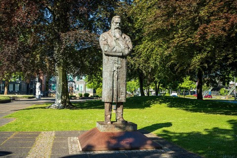 Statue of a man with crossed arms in a bright green park with trees in the background, statue of