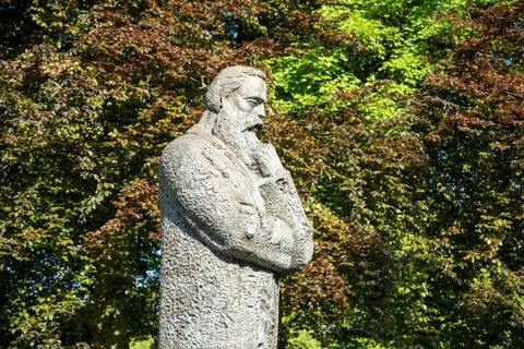 Stone statue of a pensive man surrounded by lush green trees in a park, statue of Friedrich Engels