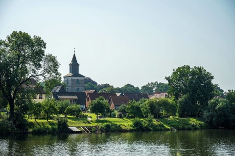 Village by the river with church and lush greenery, relaxed rural atmosphere under a clear sky,