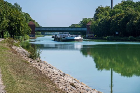 Cargo ship under a bridge, flanked by trees. Tranquil river landscape with greenery, Dortmund-Ems
