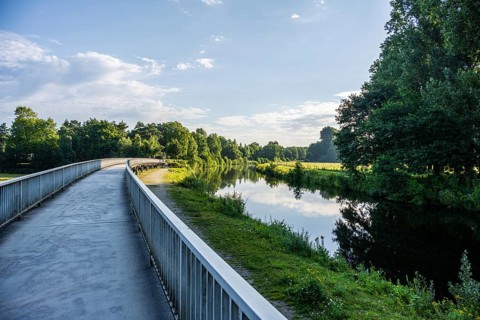 Path along a calm river, lush vegetation and cloudy sky are reflected in the water, Emssee near