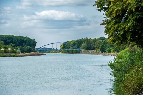 View of a canal with a modern blue bridge under a cloudy sky, surrounded by green trees, Senden,