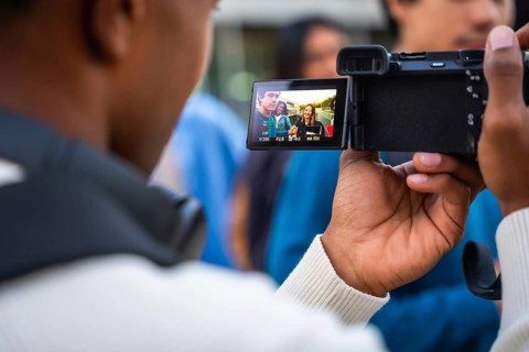 Young person holding a digital camera, filming three diverse students smiling and engaging in