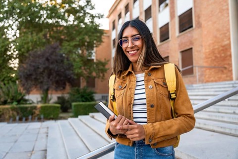 Young woman smiling, wearing glasses and a backpack, holding a book, and standing on university