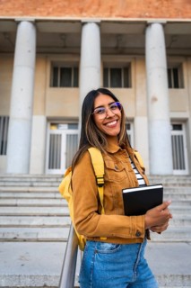 Young woman smiling, wearing glasses and a backpack, standing with a notebook in front of a