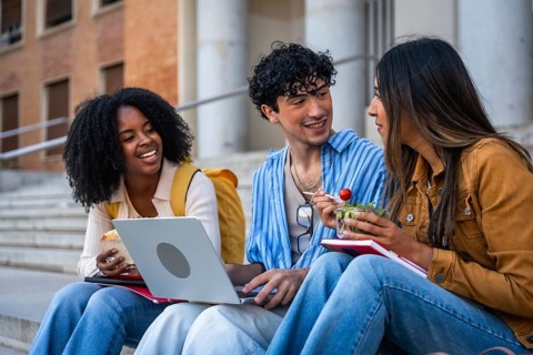 Group of multi ethnic university students sitting on outdoor steps, sharing a light meal, laughing,
