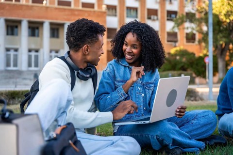 Young diverse students sitting on grass, using a laptop together and discussing ideas, reflecting
