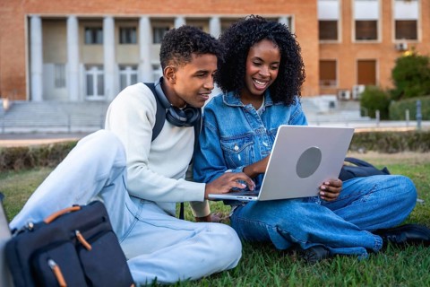 Young black students sitting on green grass, working on a laptop, discussing school project or